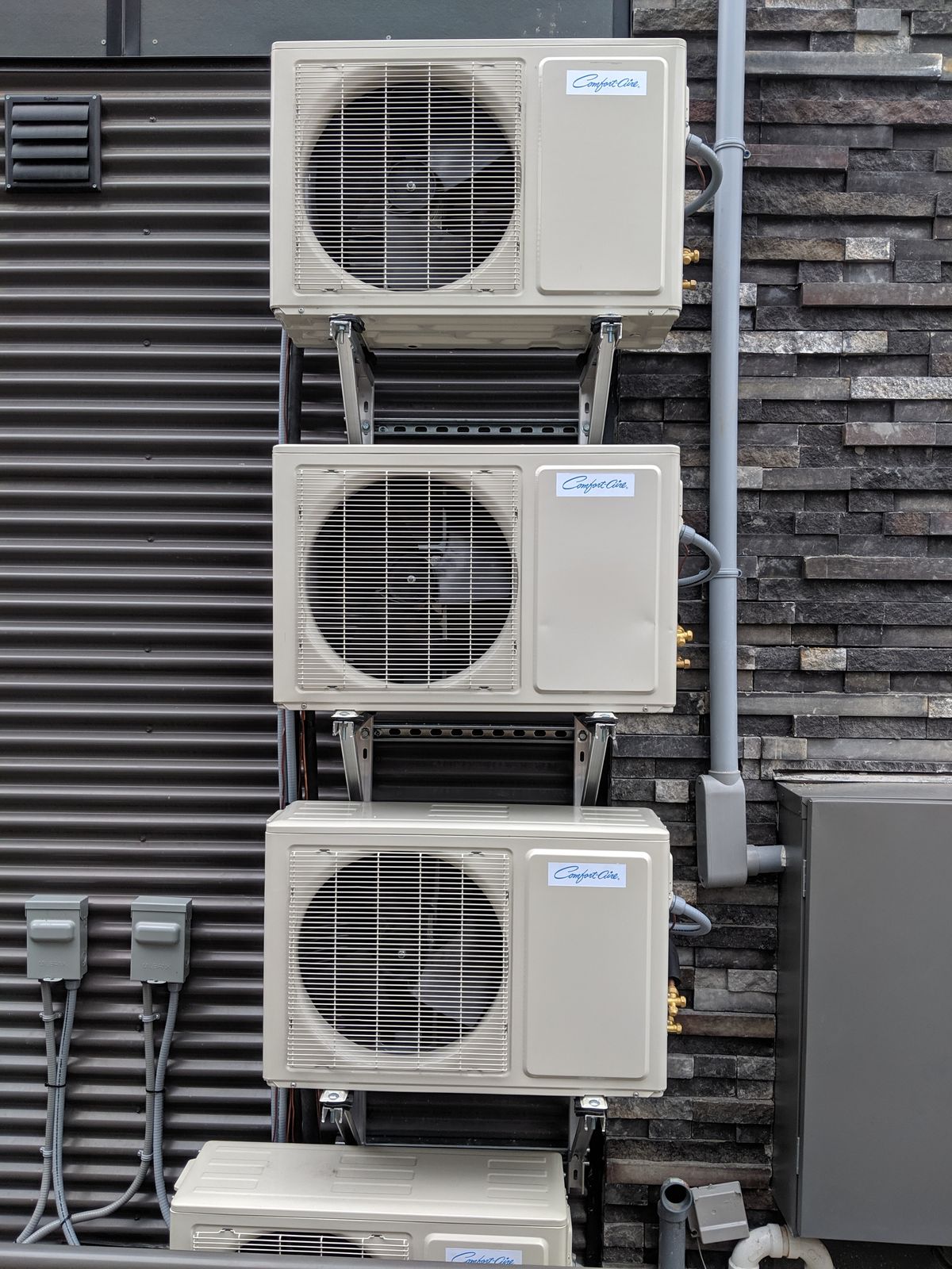 Three ductless heat pump outdoor units stacked on wall brackets against a brick wall