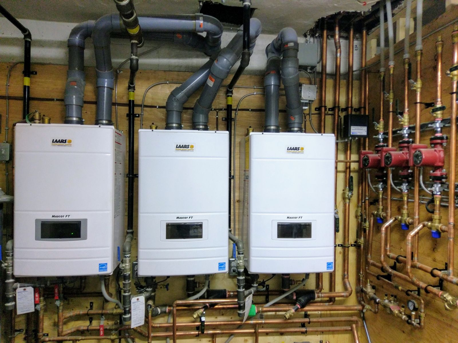 Three white wall-hung boilers with venting and copper piping on a plywood wall